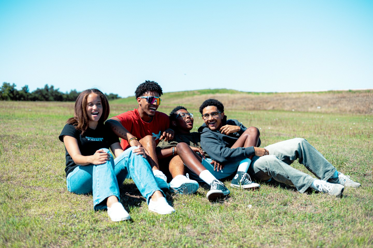 Four friends laughing together while sitting on a grassy hill.